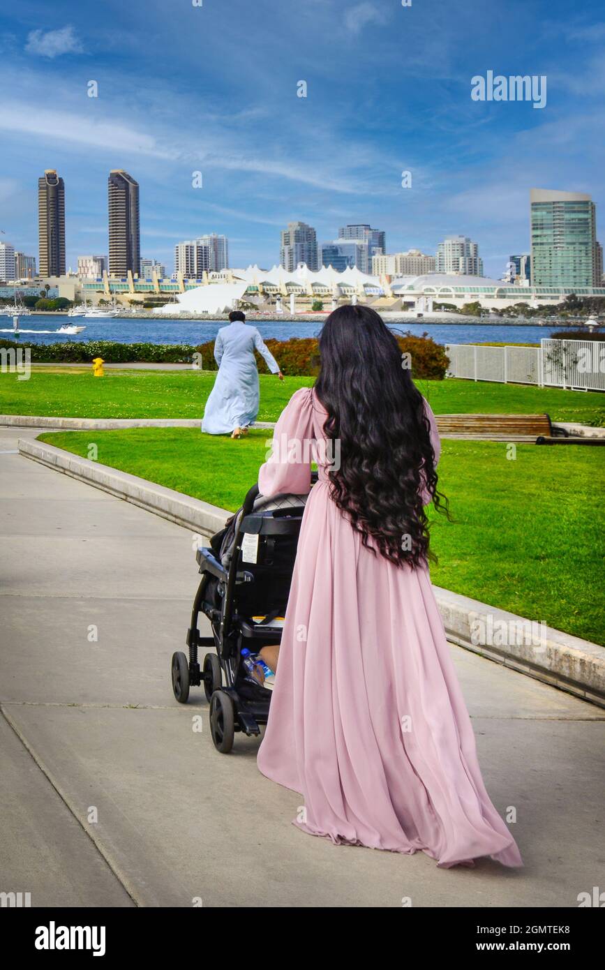 Rückansicht von Frauen mit langen schwarzen Haaren in einem langen rosa Kleid, die einen Kinderwagen auf dem Weg zur Coronado Ferry mit der Landung nach San Diego über die Bucht, CA, schieben Stockfoto