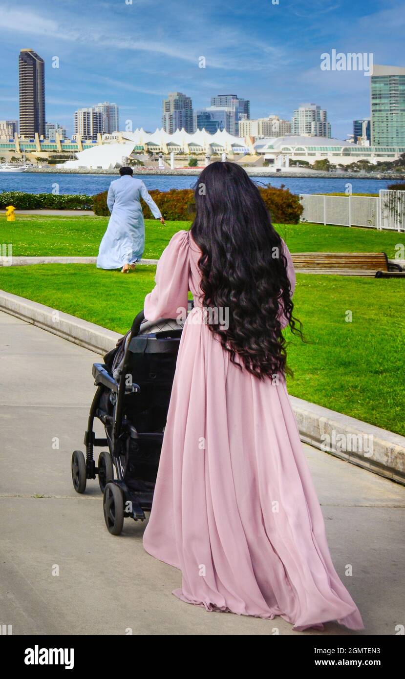 Rückansicht von Frauen mit langen schwarzen Haaren in einem langen rosa Kleid, die einen Kinderwagen auf dem Weg zur Coronado Ferry mit der Landung nach San Diego über die Bucht, CA, schieben Stockfoto