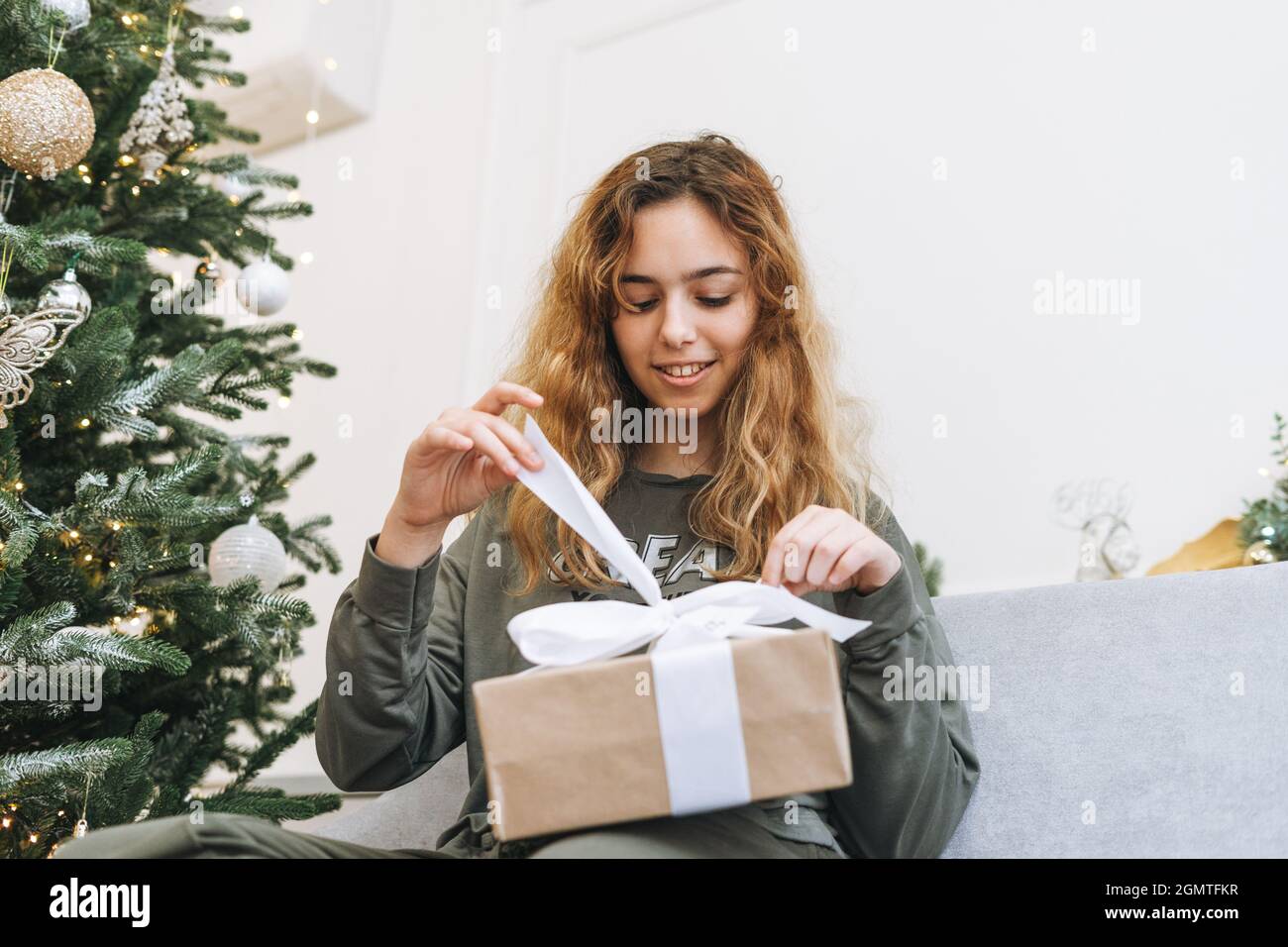 Das Mädchen im Teenageralter mit lockigen Haaren hält eine Geschenkbox in ihren Armen, während es mit einem Weihnachtsbaum auf dem Sofa im Wohnzimmer sitzt. Fröhlicher Teenager in dekorieren Stockfoto