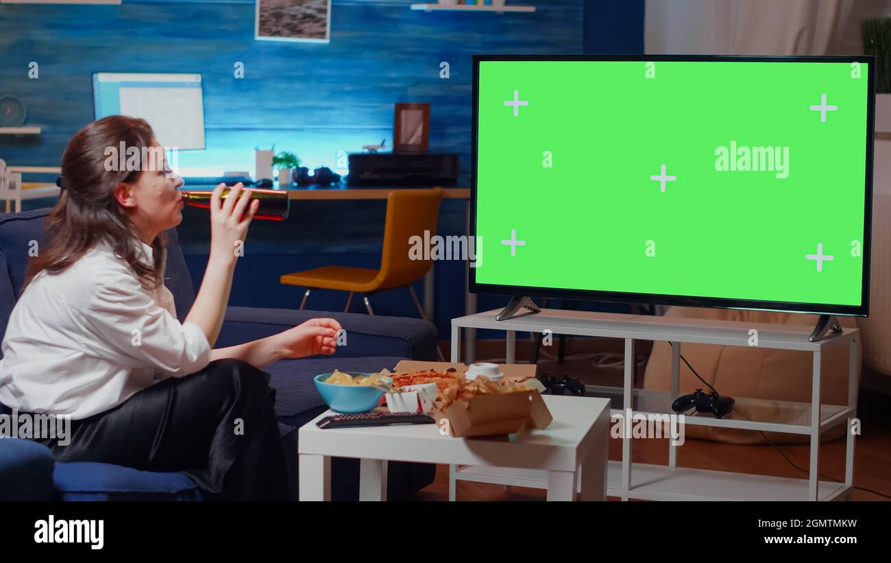 Kaukasische Frau, die beim Essen von Pommes Frites und beim Biertrinken auf dem Fernseher einen grünen Bildschirm sieht. Person, die auf der Couch sitzt und dabei die horizontale Mockup-Vorlage und den isolierten Hintergrund beobachtet, während sie lacht Stockfoto