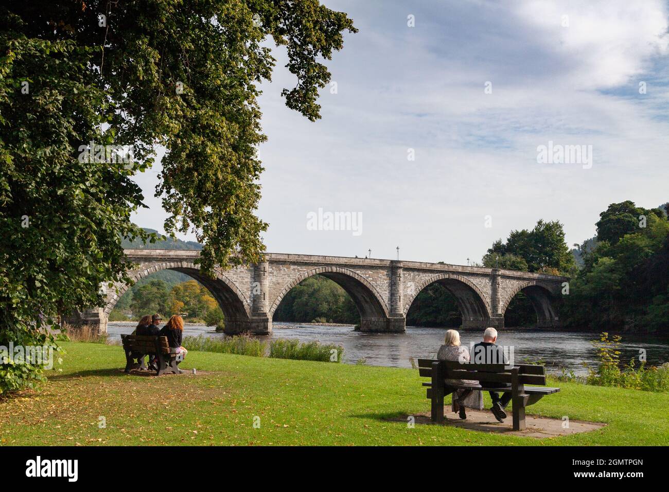 Menschen sitzen auf Bänken vor der Brücke über den Tay in Dunkeld Stockfoto