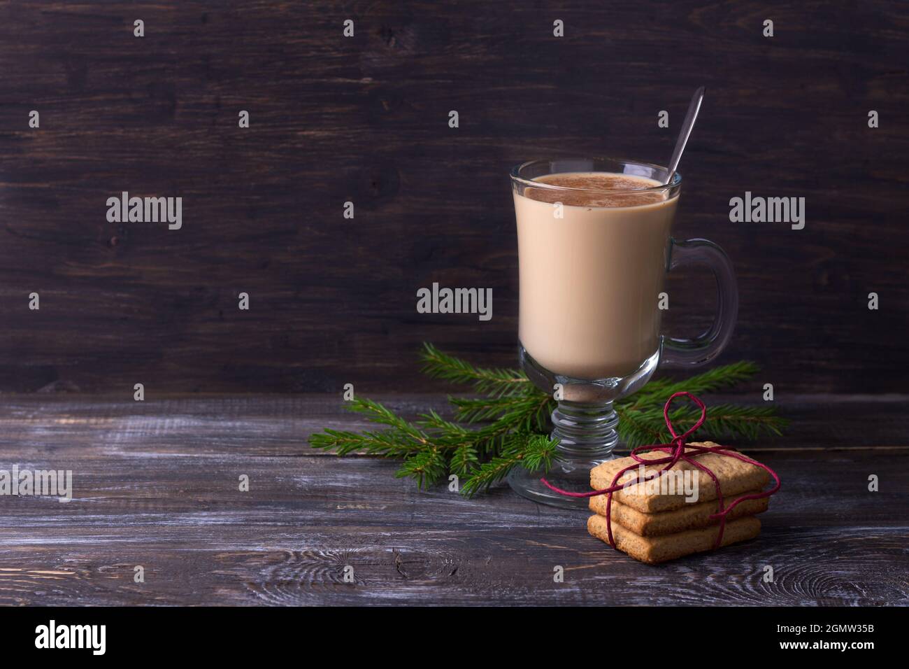 Würziger, wärmender Tee mit Milch in einer Glasschale mit Zimt, Lebkuchenkeks mit Band und Christbaumzweigen auf einem Holztisch, Freigiebel Stockfoto
