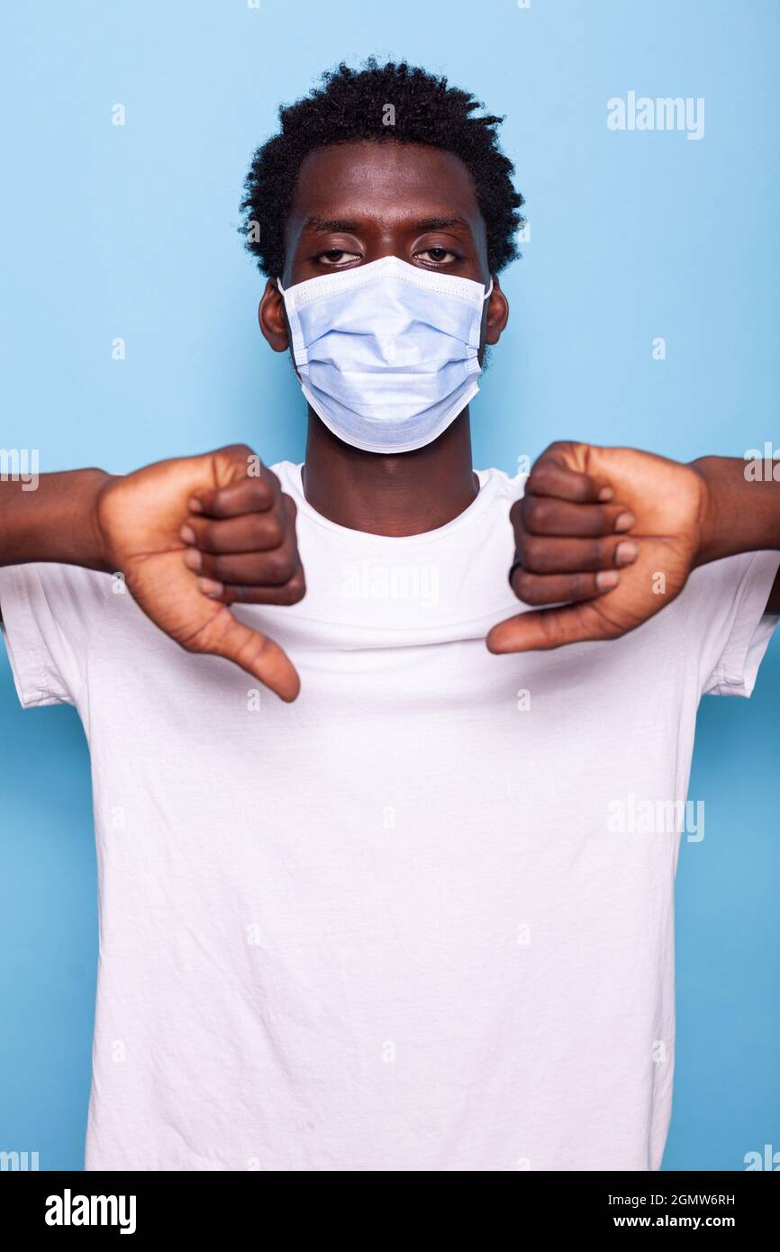 Junger Erwachsener zeigt Daumen nach unten Geste mit den Händen. Zwangloser Mann, der schlechtes Schild mit den Fingern hält, während er die Kamera anschaut und eine Gesichtsmaske trägt, um sich vor einer Coronavirus-Pandemie zu schützen. Stockfoto