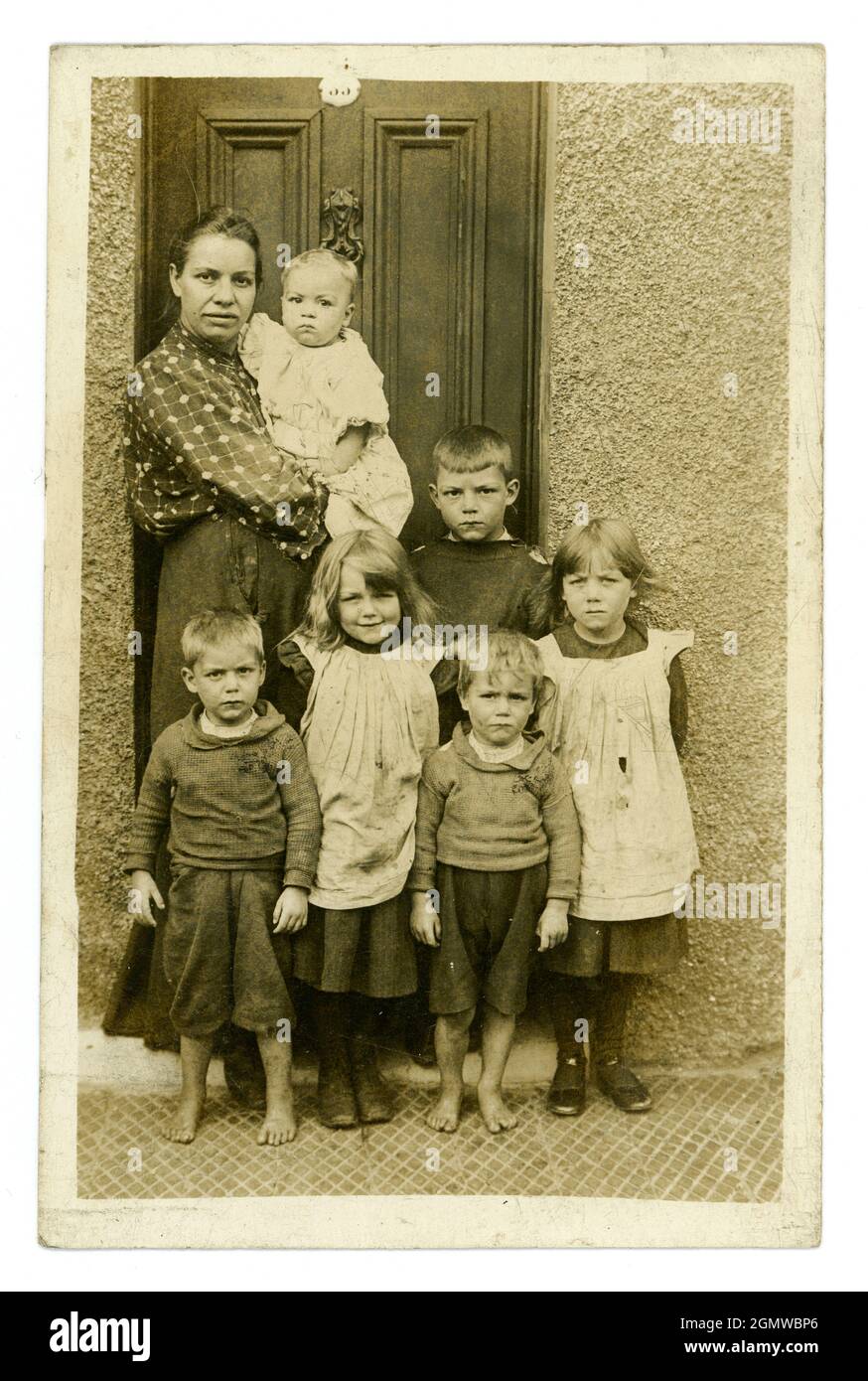 Ursprüngliche, sehr klare, edwardianische Porträtpostkarte mit einer typischen armen Arbeitermutter mit einer großen Familie von sechs ernst aussehenden kleinen Kindern und einem Baby in ihren Armen, einige Kinder sind barfuß, tragen schmuddelige, zerrissene Kleidung. Die Mädchen sind in Schürzkügelchen gekleidet, Jungen tragen knabberige Pullover, vor der Haustür, aus dem Studio von J.H. Ormerod, Seacombe, Wallasey, The Wirral, England, Großbritannien um 1907 Stockfoto