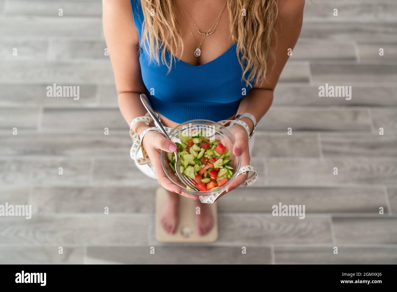 Junge Frau isst Salat für ein gesundes Leben und einen gesunden Körper. Hält ein Maßband, um ihre Taille zu messen und steht auf einer Waage. Stockfoto