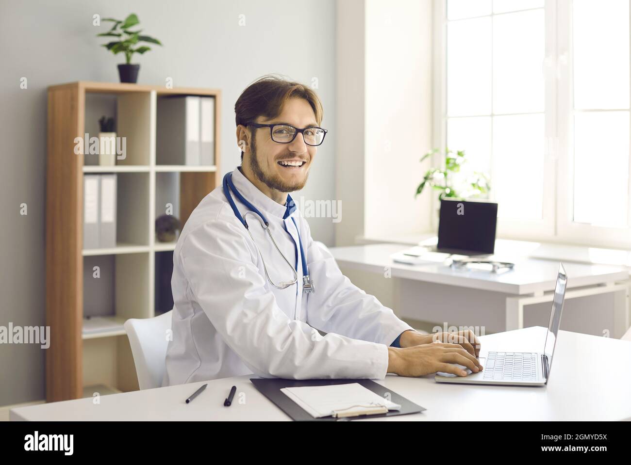 Porträt eines glücklichen jungen männlichen Arztes, der am Laptop an seinem Arbeitsplatz im Krankenhausbüro sitzt. Stockfoto