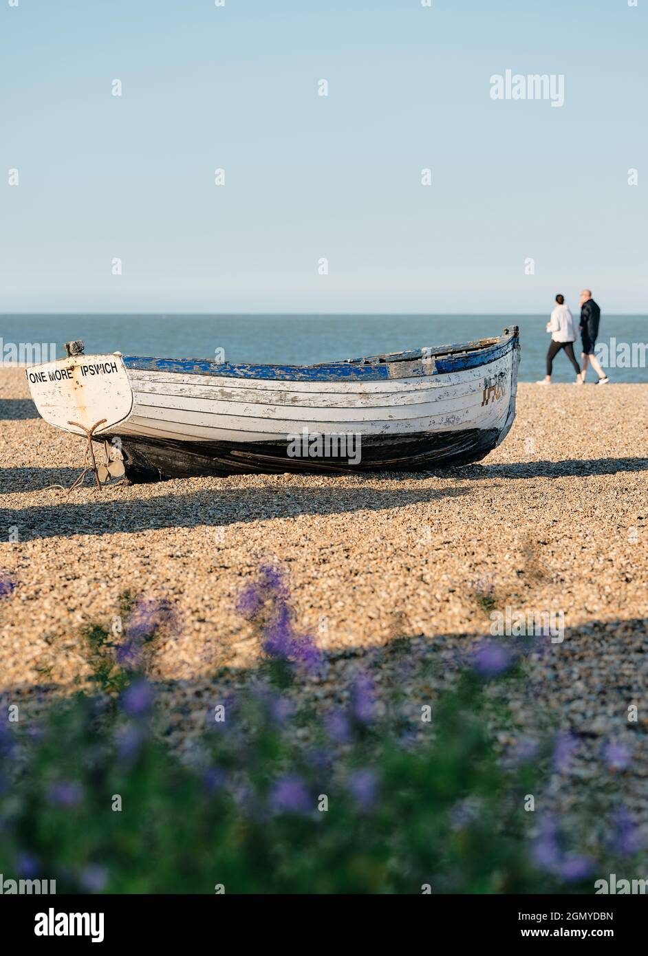 Ein altes Fischerboot und ein Touristenpaar, das am Sommerstrand in der Abendsonne an der Aldeburgh Suffolk Coast England UK spazieren geht - Aufenthalt Sommerurlaub Stockfoto