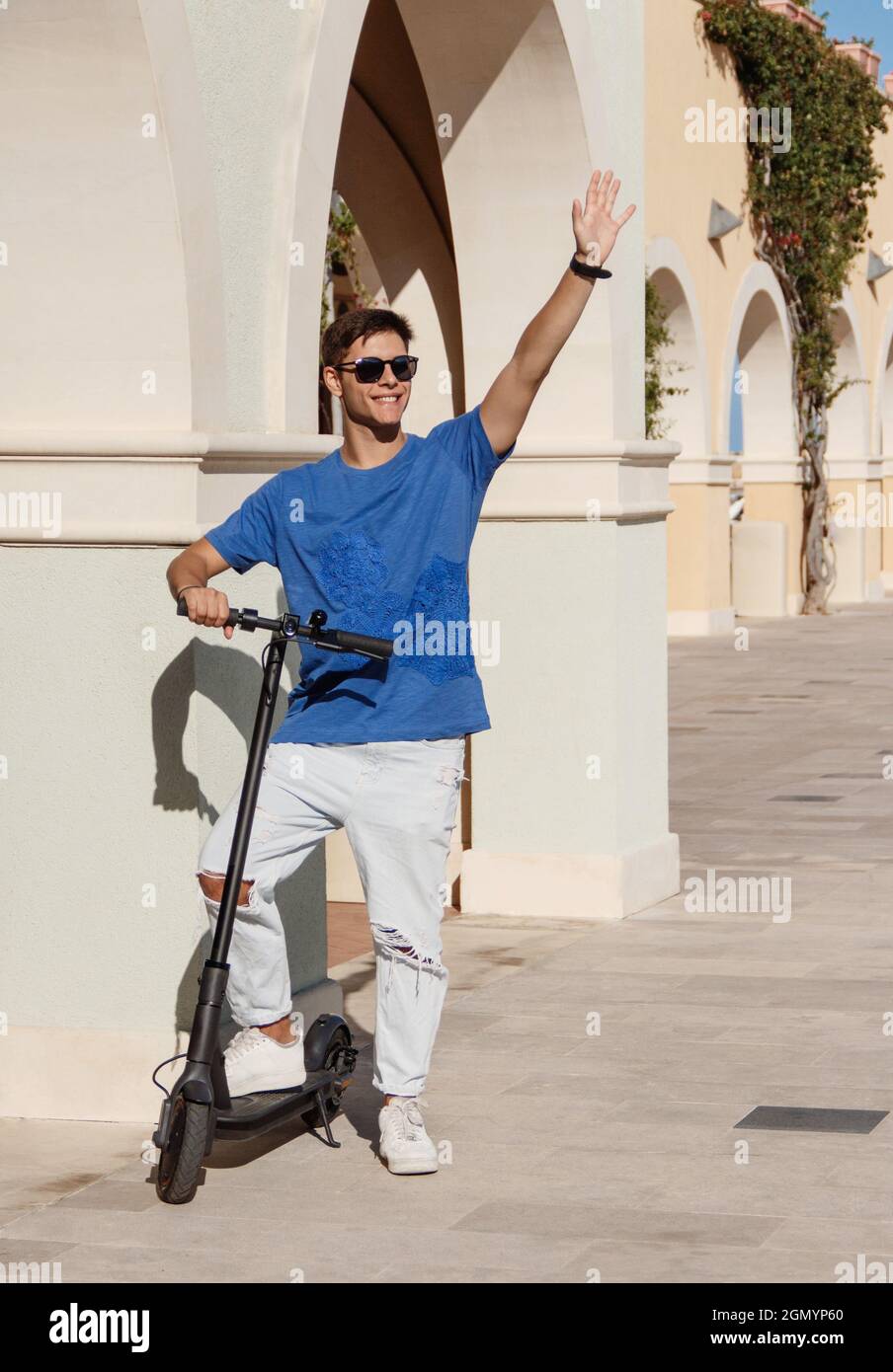 Stadtverkehr. Glücklicher junger Mann in Freizeitkleidung mit Elektro-Push-Scooter auf der Stadtstraße, winkt zu jemandem, Gruß Freund im Freien, kopieren Raum. A Stockfoto