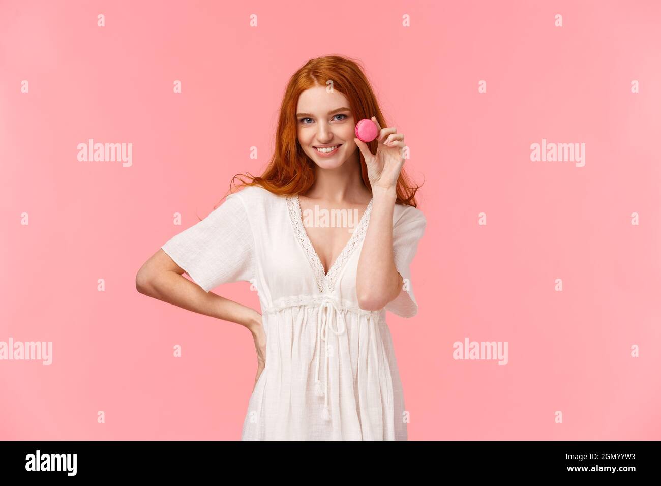 Schönheit, Desserts und Personen Konzept. Betörend und sassy redhead kaukasische Mädchen in weißem Kleid, jemand verführt mit lecker, lecker Macaron, lächelnd Stockfoto