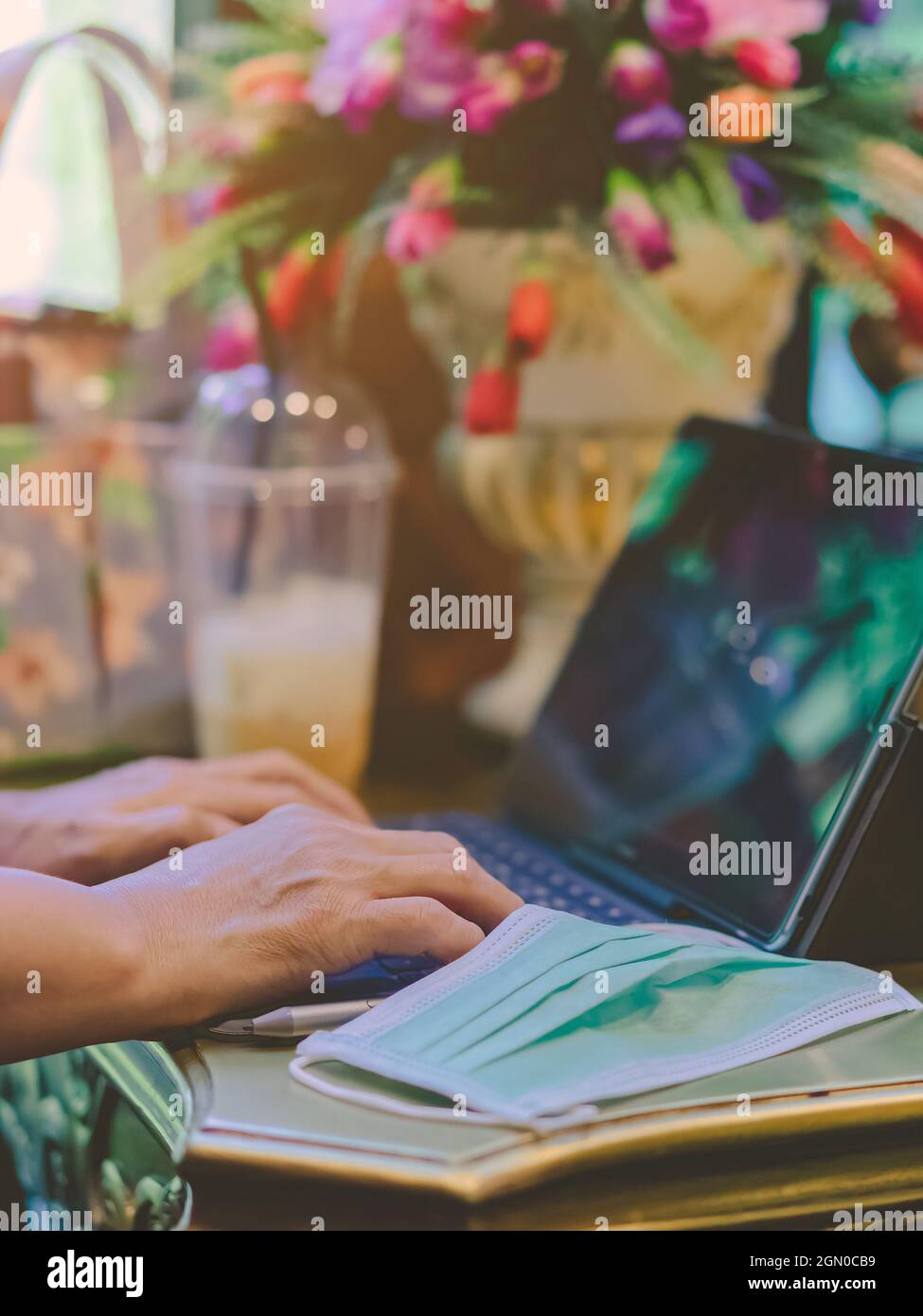 Hände einer reifen Frau, die auf der Laptop-Tastatur tippt, um von zu Hause aus zu arbeiten und ihre OP-Maske auszuziehen, legen sie auf den Tisch, damit sie leichter atmen kann. Konzepte von C Stockfoto