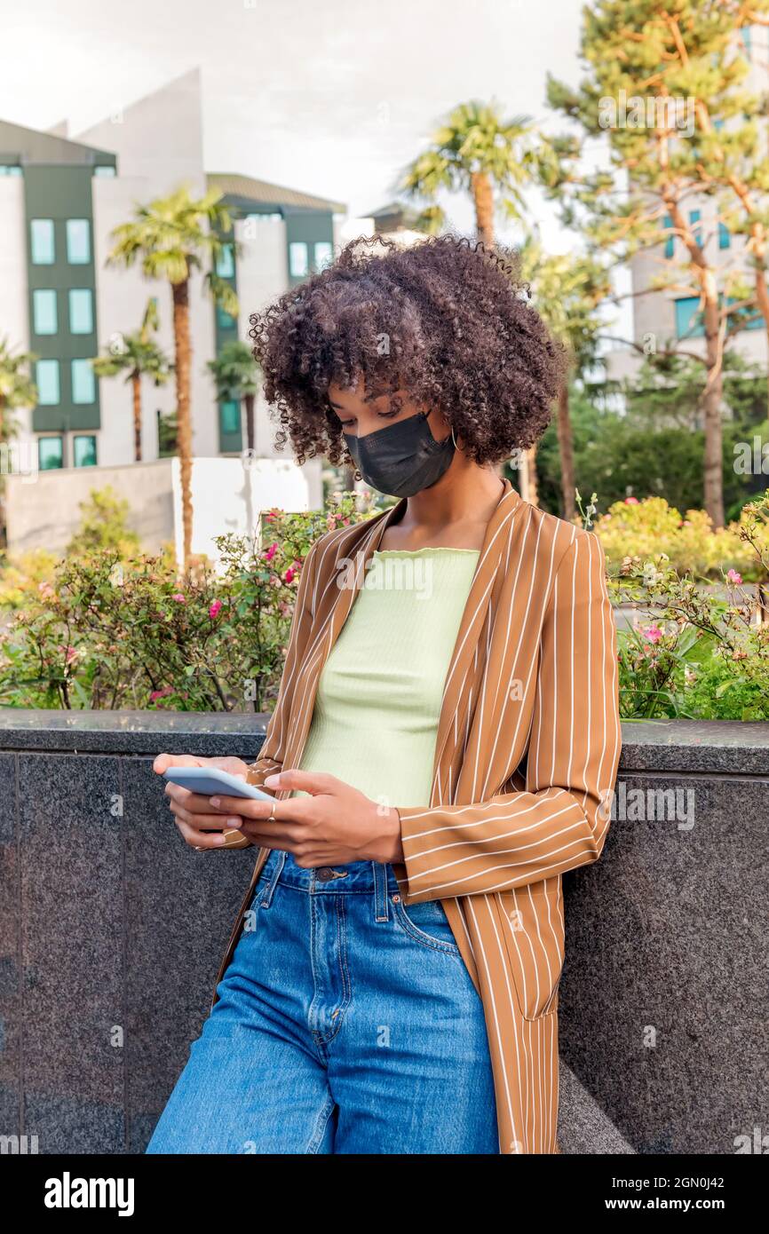 Seriöse afroamerikanische Frau mit Afro-Frisur und in Schutzmaske, die auf der Straße steht und während des Coronavirus auf dem Mobiltelefon Nachrichten ausgibt Stockfoto