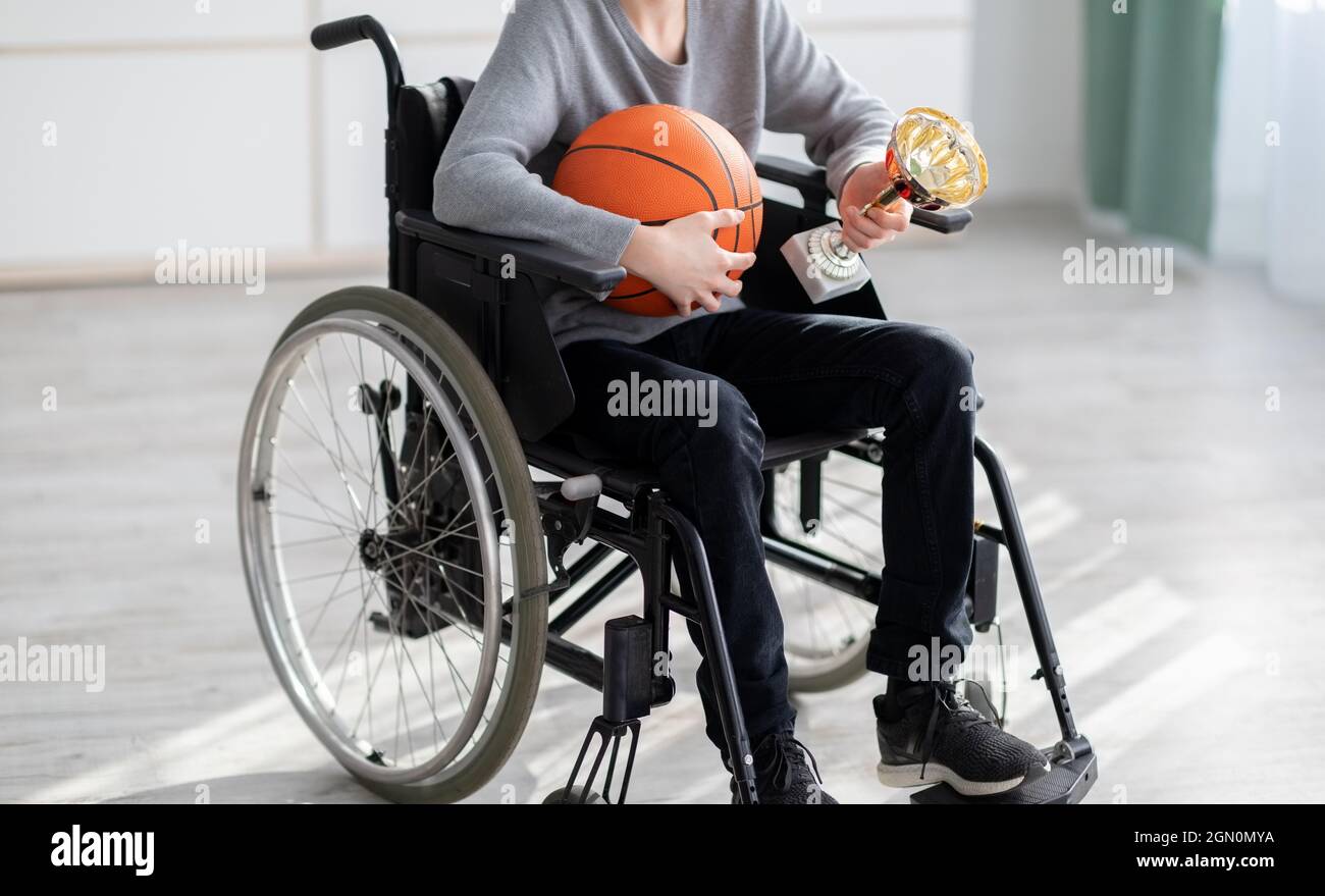 Nicht erkennbarer Teenager-Basketballspieler mit Ball und Trophäe sitzt im Rollstuhl und leidet wegen Verletzungen zu Hause Stockfoto