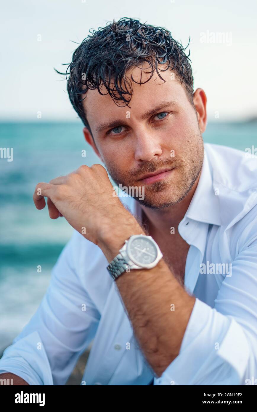 Ernsthafter Rüde mit nassem Haar und in weißem Hemd, der auf die Kamera auf dem Meeresgrund am Strand schaut Stockfoto