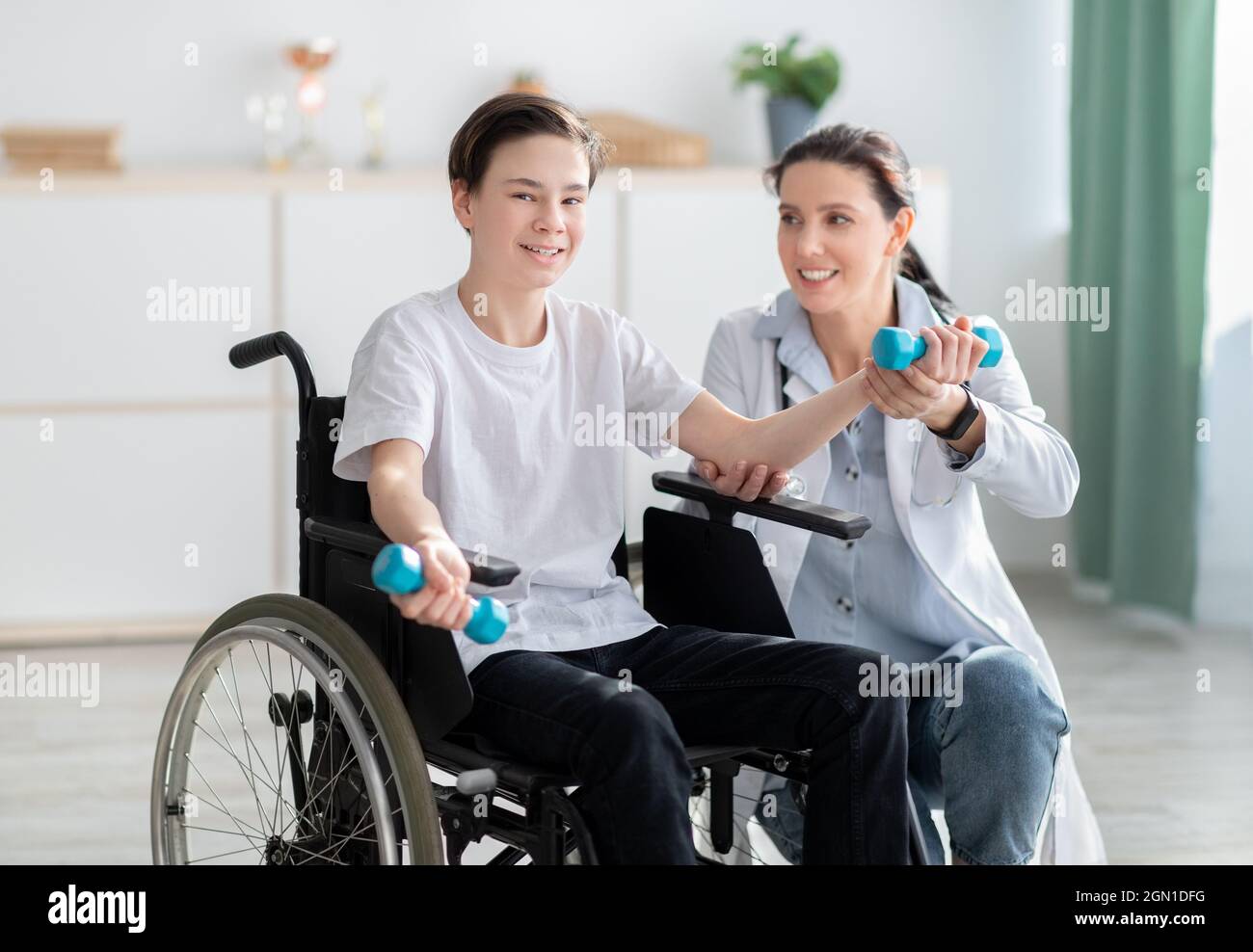 Physiotherapeutin, die einem glücklichen Teenager im Rollstuhl hilft, zu Hause Übungen zu machen. Körperliche Rehabilitation Stockfoto