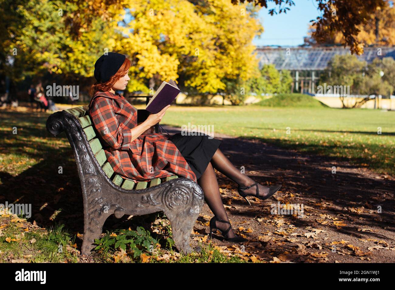 Rothaarige Frau im karierten Mantel und schwarzer Baskenmütze liest ein Buch auf der Bank und ruht sich an sonnigen Tagen im Herbstpark aus Stockfoto