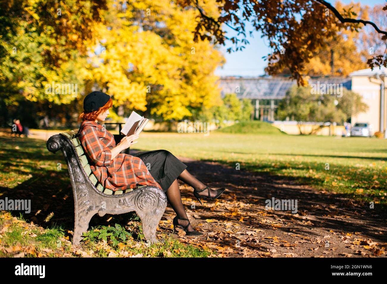 Rothaarige Frau im karierten Mantel und schwarzer Baskenmütze liest ein Buch auf der Bank und ruht sich an sonnigen Tagen im Herbstpark aus Stockfoto