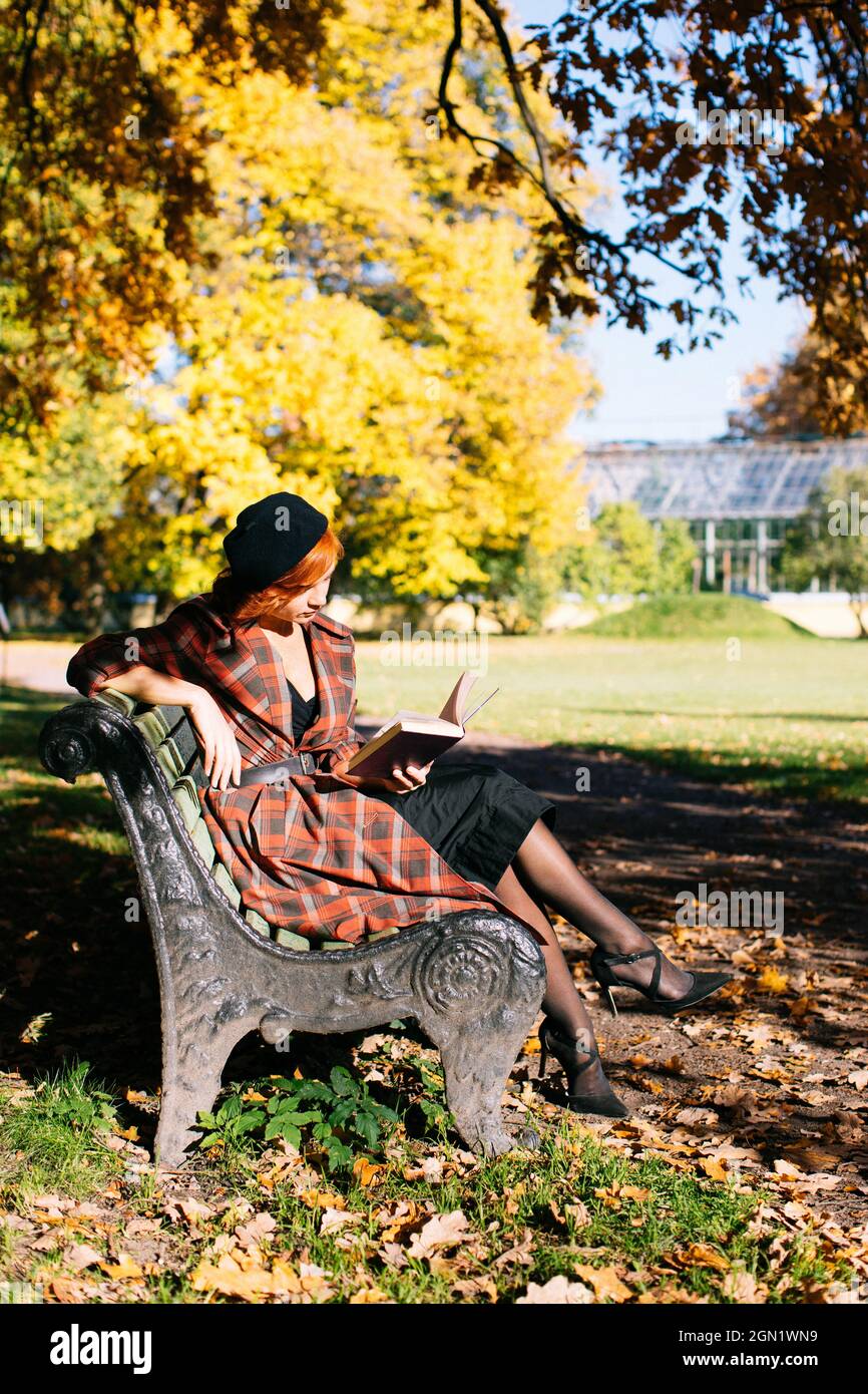 Rothaarige Frau in kariertem Mantel und schwarzem Baskenmütze, die auf der Bank liest und sich an sonnigen Tagen im Herbstpark ausruht Stockfoto