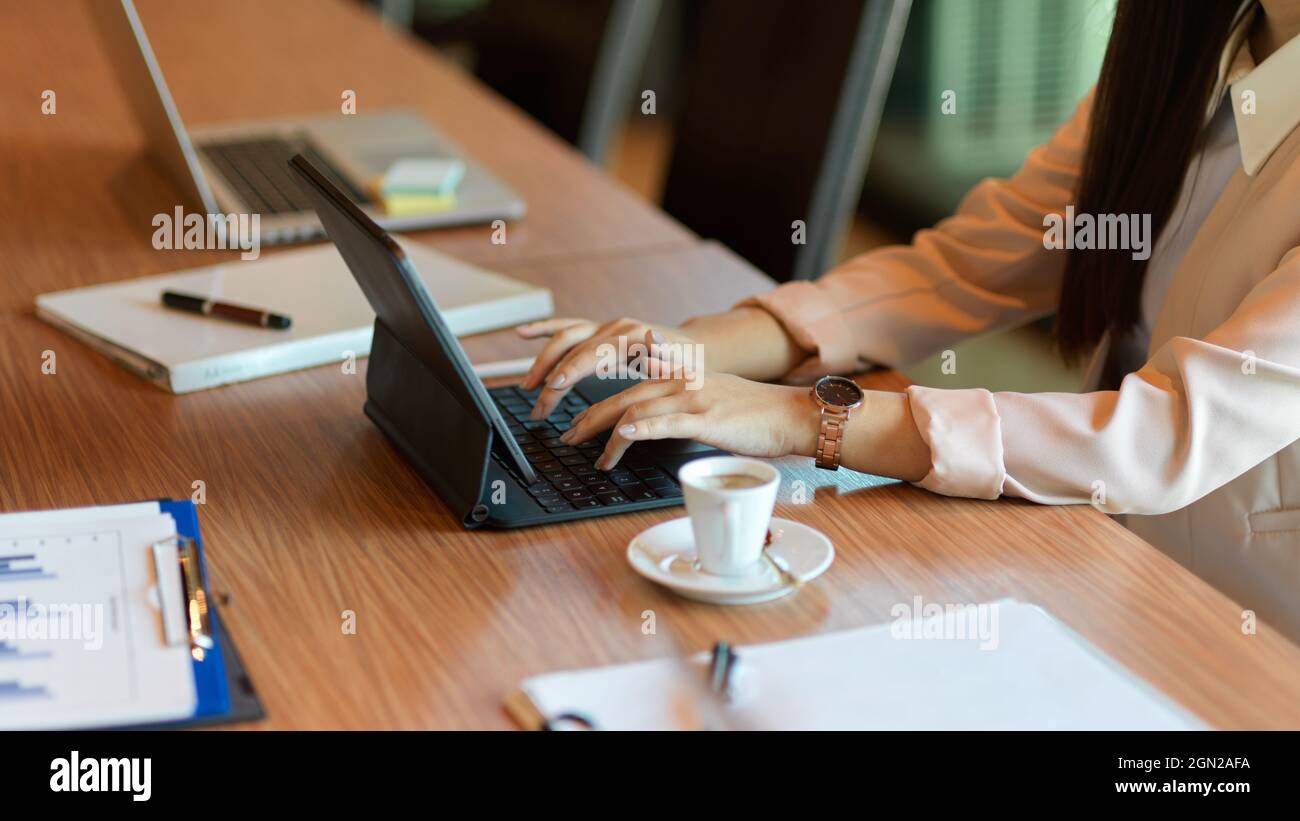 Zugeschnittenes Bild einer Geschäftsfrau, die auf einem tragbaren Tablet arbeitet, einer Assistentin, die auf der Tastatur tippt, während sich das Unternehmen in einem Besprechungsraum trifft. Stockfoto