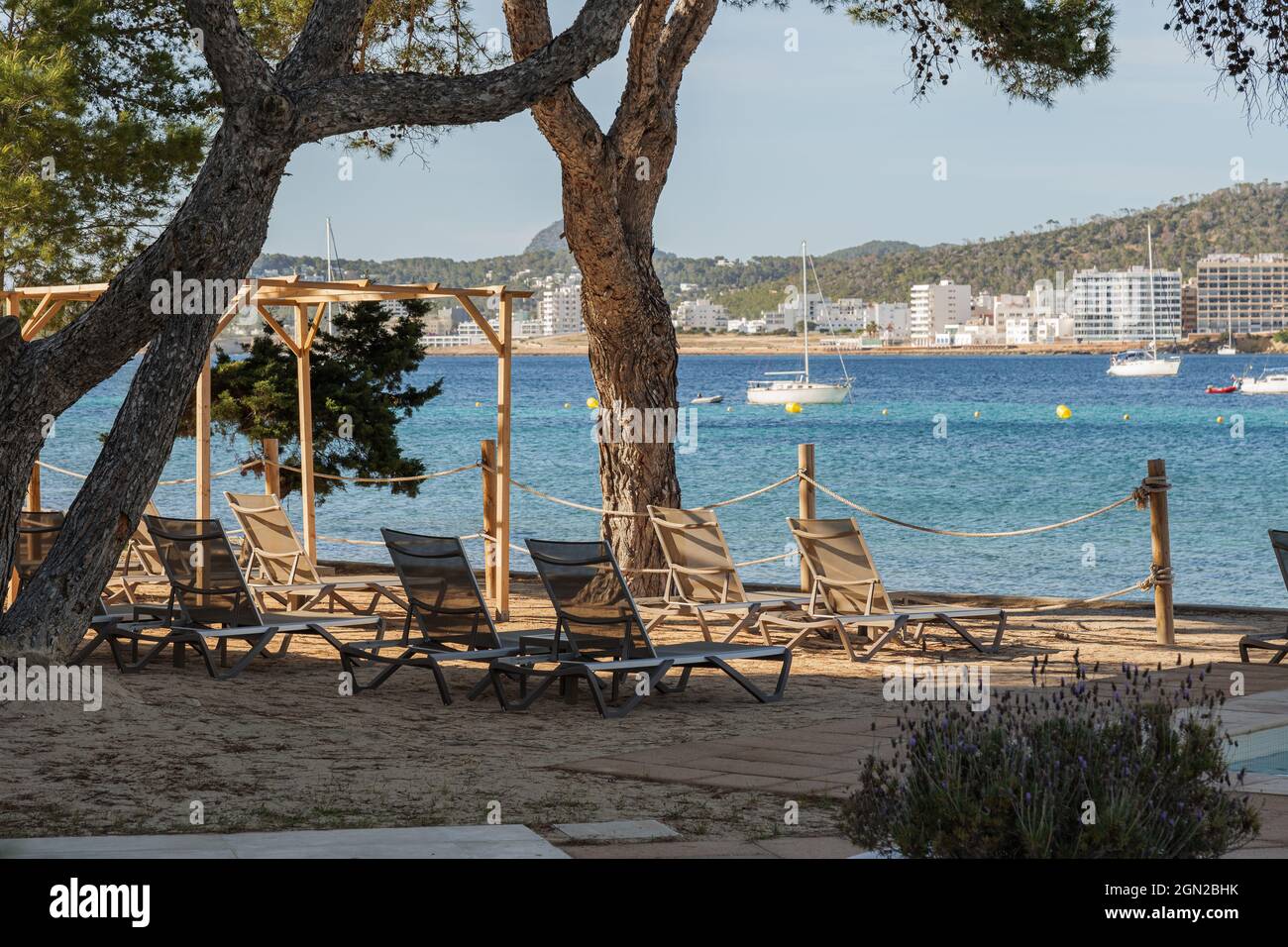 Sonnenliegen am Strand mit Blick auf eine Bucht mit Yachten auf der Insel Ibiza. Schöne Relaxzone in der Nähe des Meeres mit Liegestühlen unter großen Bäumen. Stockfoto