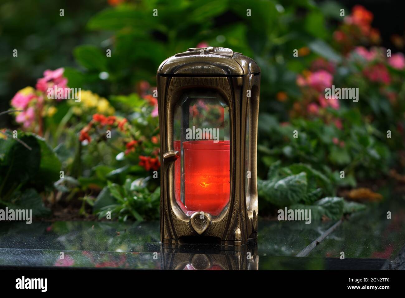 Grablampe aus Metall mit brennender Kerze auf einem Grab mit bunten Blumen in verschwommenem Hintergrund Stockfoto