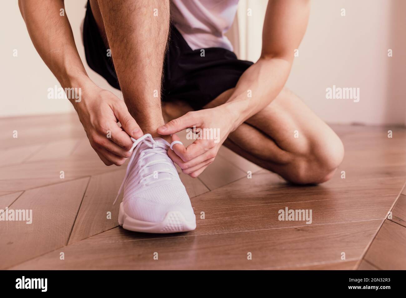 Morgendliche Trainingsroutine im Fitnessstudio zu Hause. Fitness Motivation und Muskeltraining Konzept. Mann in weißen Sneakers, der Schnürsenkel im Sonnenlicht bindet Stockfoto