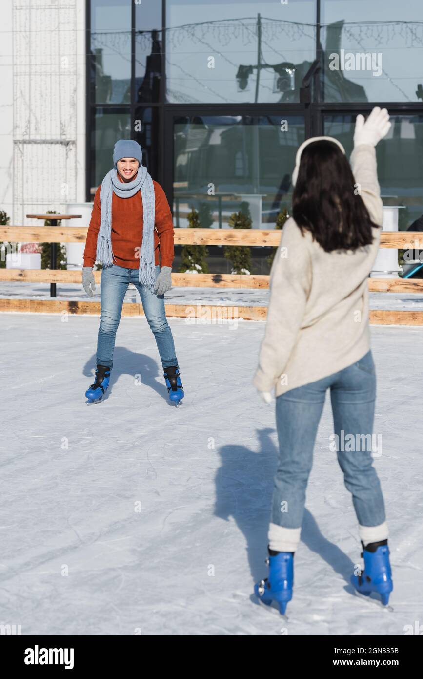 In voller Länge glücklicher Mann im Hut Blick auf verschwommene Frau winken Hand auf Eisbahn Stockfoto