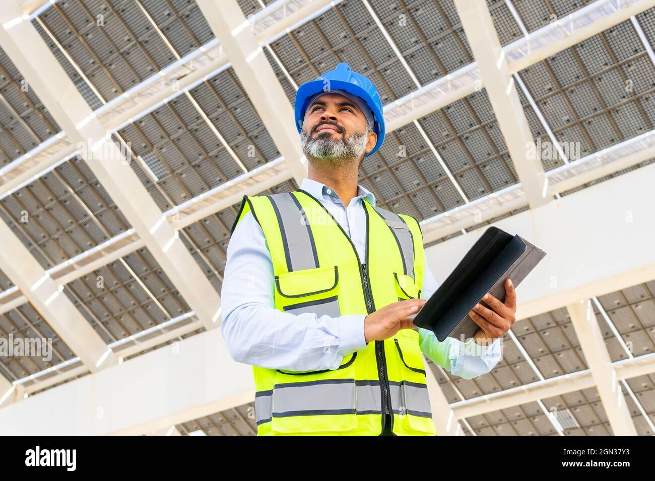 Niedriger Winkel eines männlichen hispanischen Ingenieurs mittleren Alters in Uniform mit Tablet, das unter dem Solarkraftwerk wegschaut Stockfoto