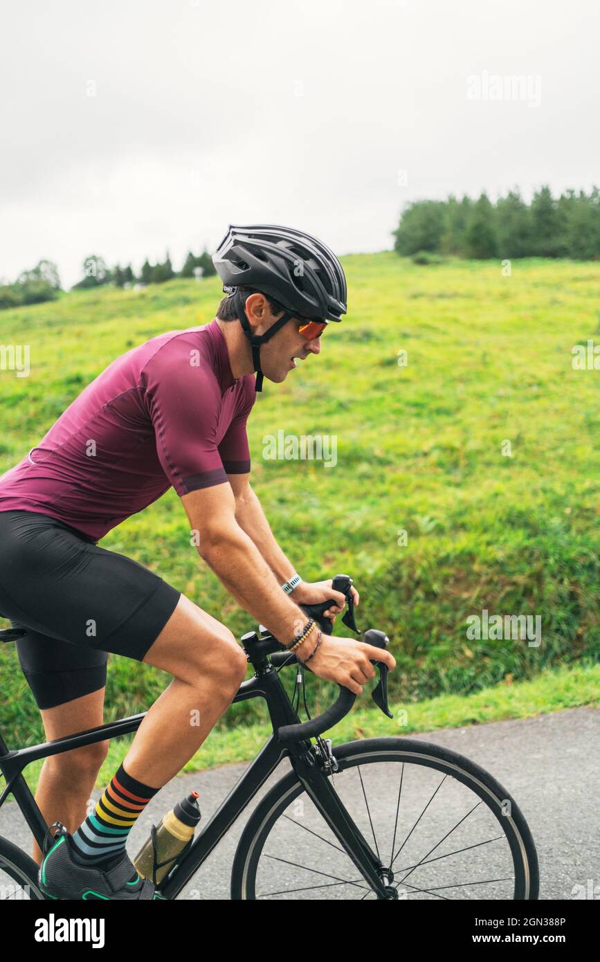 Seitenansicht des Sportlers im Schutzhelm, der während des Trainings auf einer asphaltierten Straße gegen grünen Hügel und Bäume unter hellem Himmel Fahrrad fährt Stockfoto