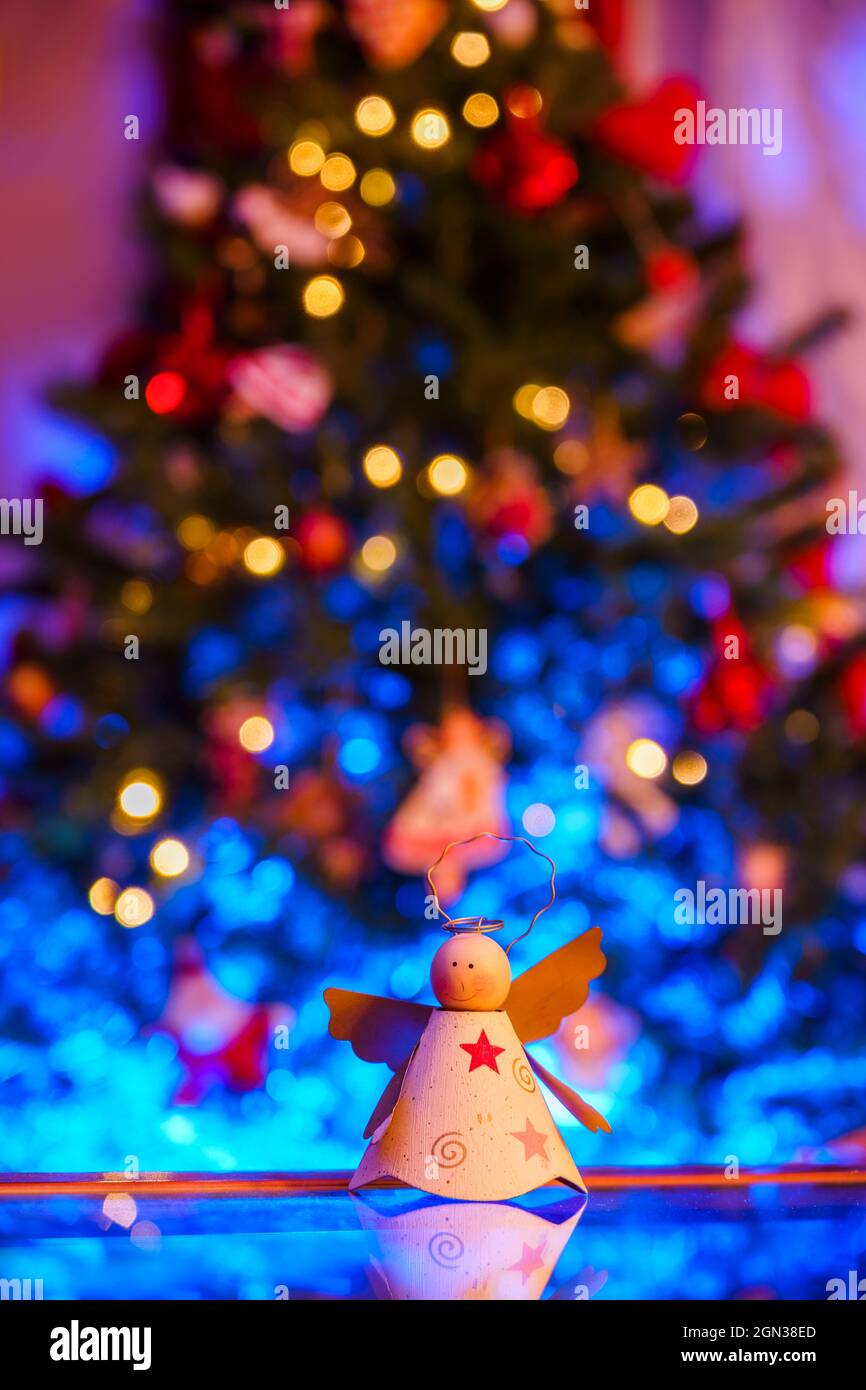 Handgemachtes, engelförmiges Spielzeug, das auf einem reflektierenden Tisch gegen einen festlichen Weihnachtsbaum mit leuchtenden Girlanden aufgestellt wird Stockfoto