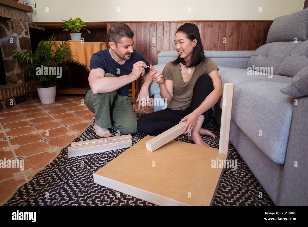 Mann, der eine Schraube an eine fröhliche asiatische Frau hergab, während er im Wohnzimmer einen Tisch auf einem Zierteppich montierete Stockfoto