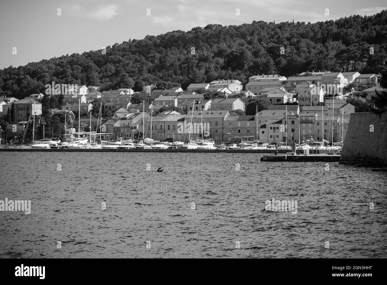 Graustufenaufnahme von Gebäuden am Wasser und einem Hafen auf dem Hügel am azurblauen See in Korcula, Kroatien Stockfoto