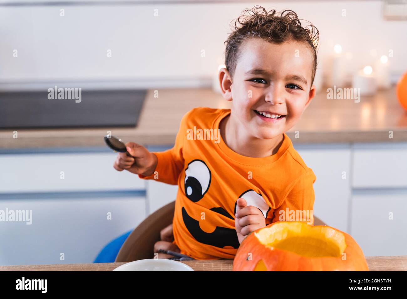 Porträt eines Jungen glücklich Blick auf Kamera gekleidet wie ein halloween Kürbis T-Shirt Entleeren und Dekorieren eines halloween Kürbis in der Küche zu Hause. Stockfoto