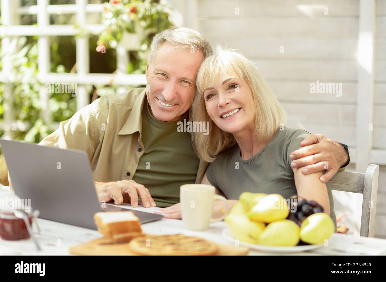 Glückliches Seniorenpaar, das im Freien einen Laptop benutzt, auf der Terrasse sitzt und vor der Kamera lächelt Stockfoto