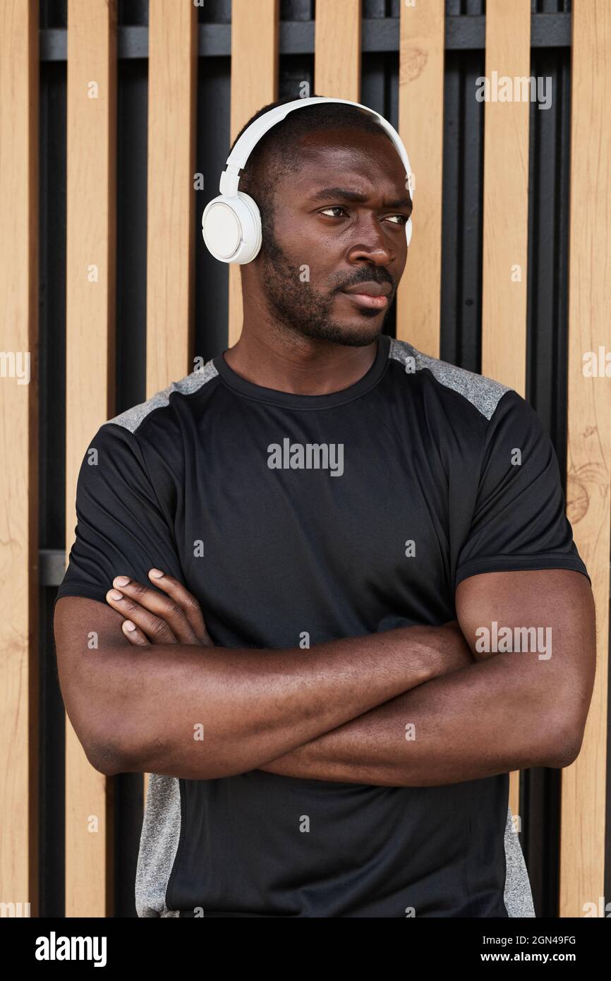 Porträt eines afrikanischen, muskulösen Mannes, der mit gekreuzten Armen im Freien steht und Musik in kabellosen Kopfhörern hört Stockfoto