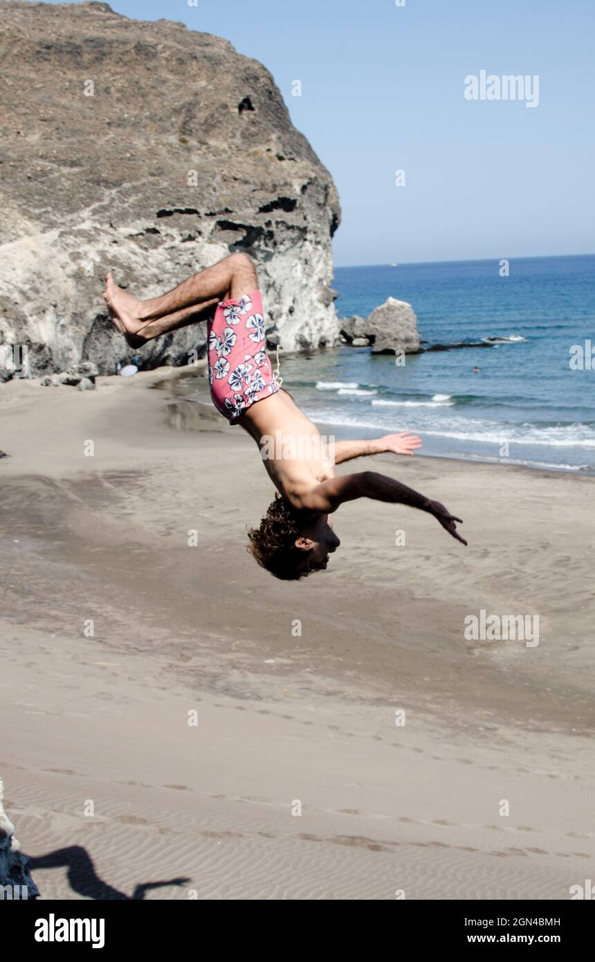 Mann im Badeanzug, der in einer Strand- und Sandlandschaft wilde Salto macht, steht auf dem Kopf in der Luft Stockfoto