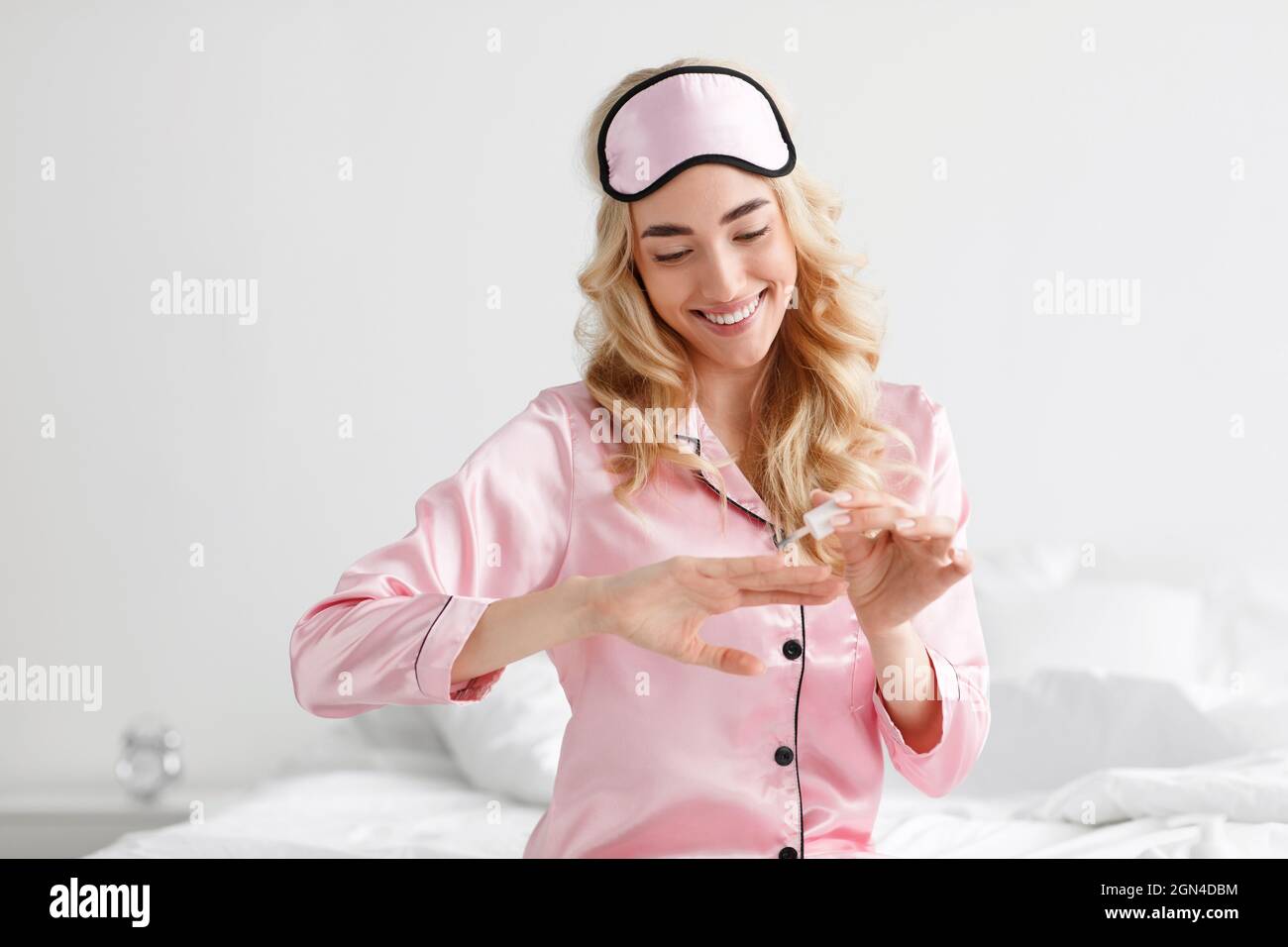 Fröhlich attraktive kaukasische blonde Dame in rosa Schlafanzug mit Schlafanzug Maske auf Nagellack gesetzt sitzt auf dem Bett Stockfoto
