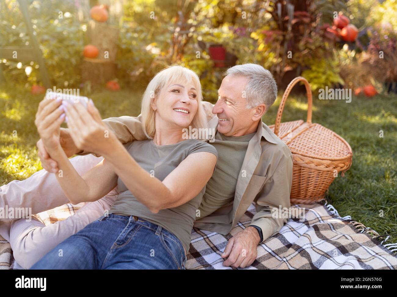 Moderne ältere Ehepartner, die Selfie auf dem Smartphone machen, Picknick machen und sich am sonnigen Herbsttag im Garten ausruhen Stockfoto