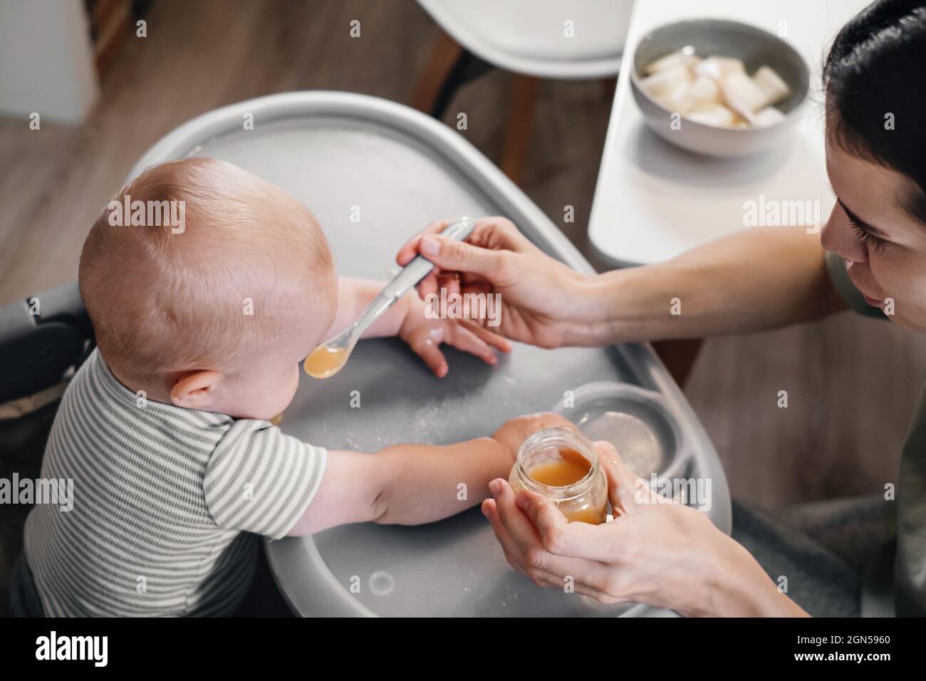 Mutter gibt dem Jungen Nahrung, rein vom Löffel im Kinderstuhl zu Hause. Ergänzende Säuglingsernährung. Glückliches Kind, das auf das Essen wartet und lernt, gerötet zu essen Stockfoto