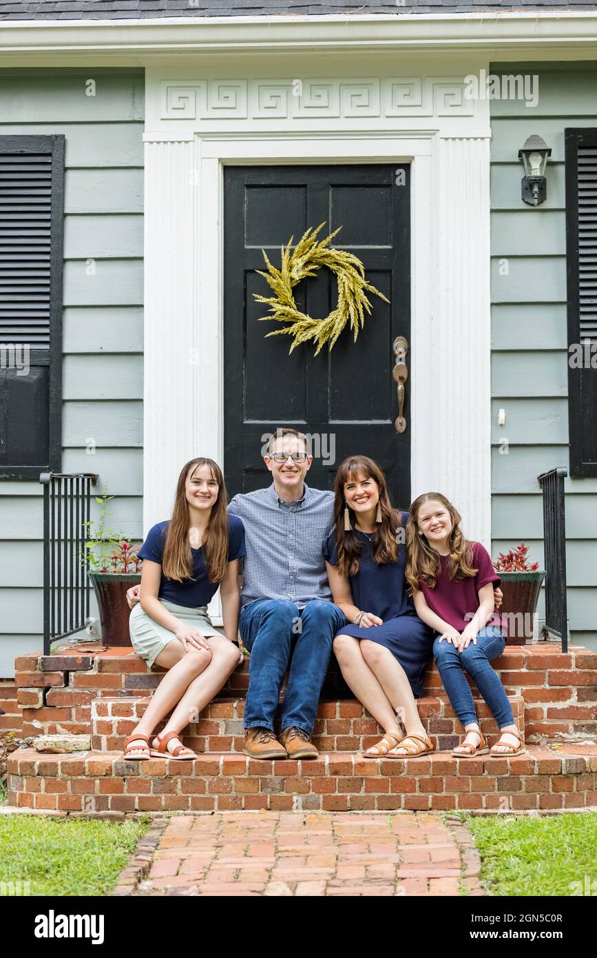 Eine Familie mit einer Mutter, einem Vater und zwei Töchtern, die draußen auf der Backsteinvertei eines kleinen blauen Häuschens sitzen Stockfoto