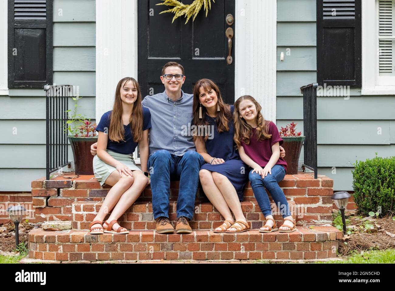 Eine Familie mit einer Mutter, einem Vater und zwei Töchtern, die draußen auf der Backsteinvertei eines kleinen blauen Häuschens sitzen Stockfoto