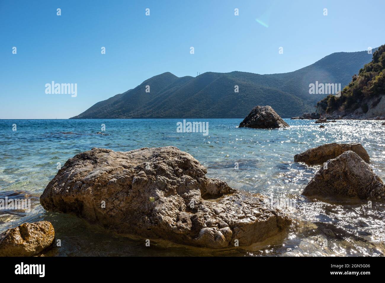 Große Felsen in kristallklarem azurblauem Wasser am sonnigen Kiesstrand mit grünen Hügeln am Horizont. Küste der Insel Lefkada in Griechenland. Sommer wilde Natur reisen Stockfoto