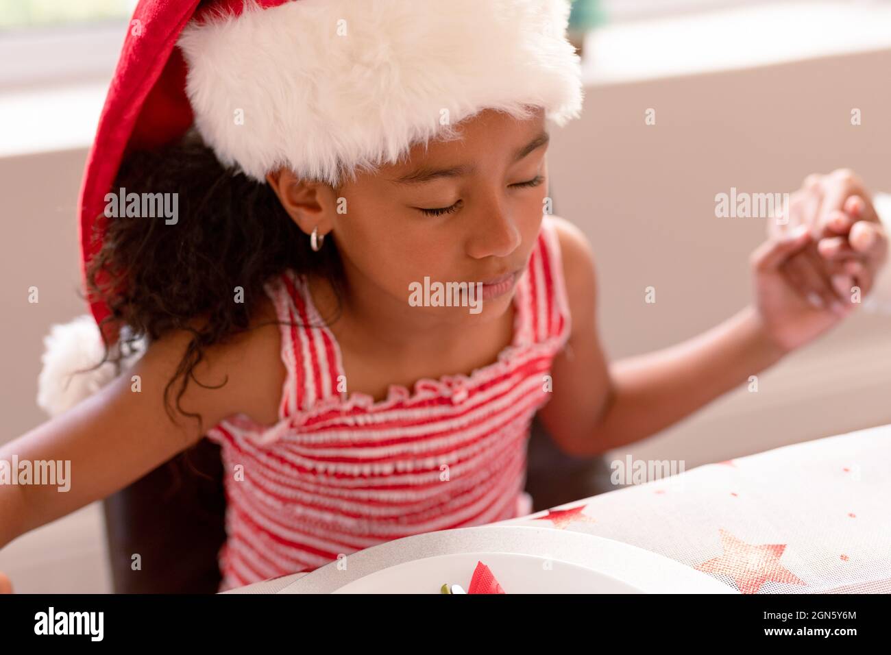 afroamerikanisches Mädchen trägt weihnachtsmütze und betet am weihnachtstisch Stockfoto