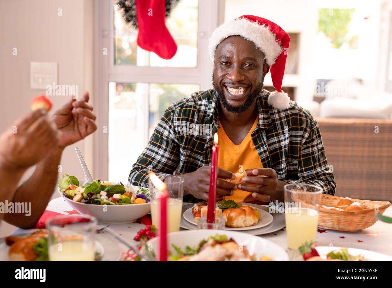 Glücklicher afroamerikanischer Mann mit weihnachtsmütze, der am weihnachtstisch sitzt Stockfoto