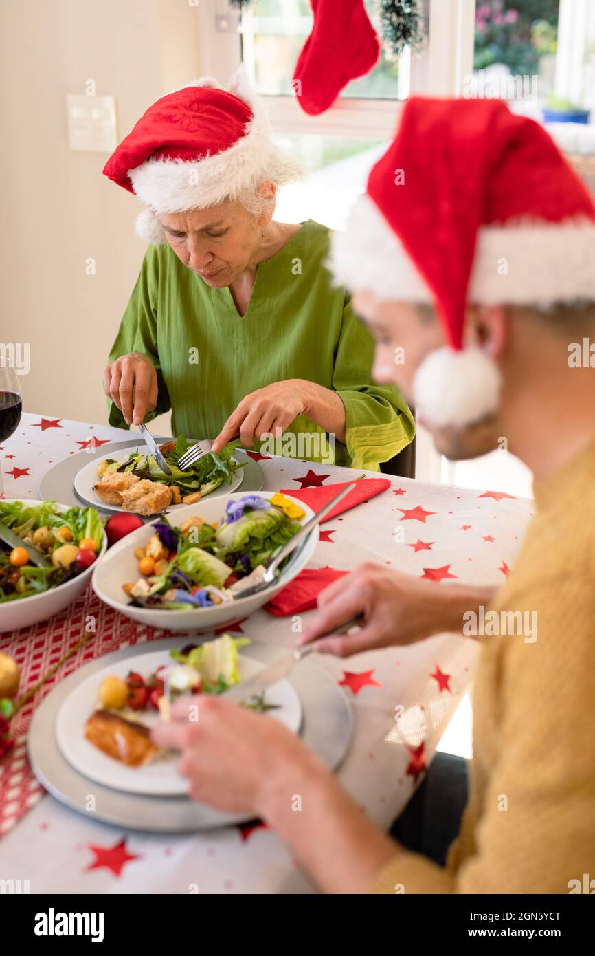 Schwiegermutter und Schwiegersohn tragen weihnachtsmützen, sitzen am weihnachtstisch und essen Stockfoto