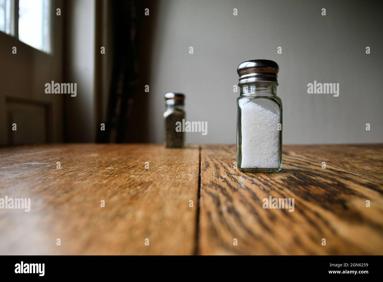 Salz- und Pfefferstreuer auf einer polierten Holzoberfläche mit Licht aus einem Fenster, das den Tisch beleuchtet, gemahlen; rustikaler Esstisch mit Glasstreuern. Stockfoto