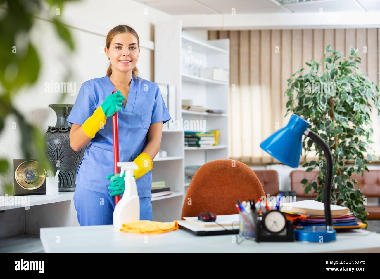 Lächelnde junge Putzfrau in Uniform, bereit für die Reinigung im Büro Stockfoto