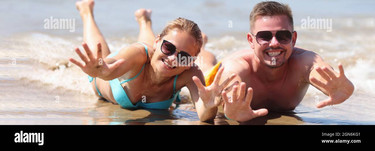 Fröhlicher Mann und Frau in Sonnenbrillen liegen auf nassem Sand In der Nähe des Meeres Stockfoto