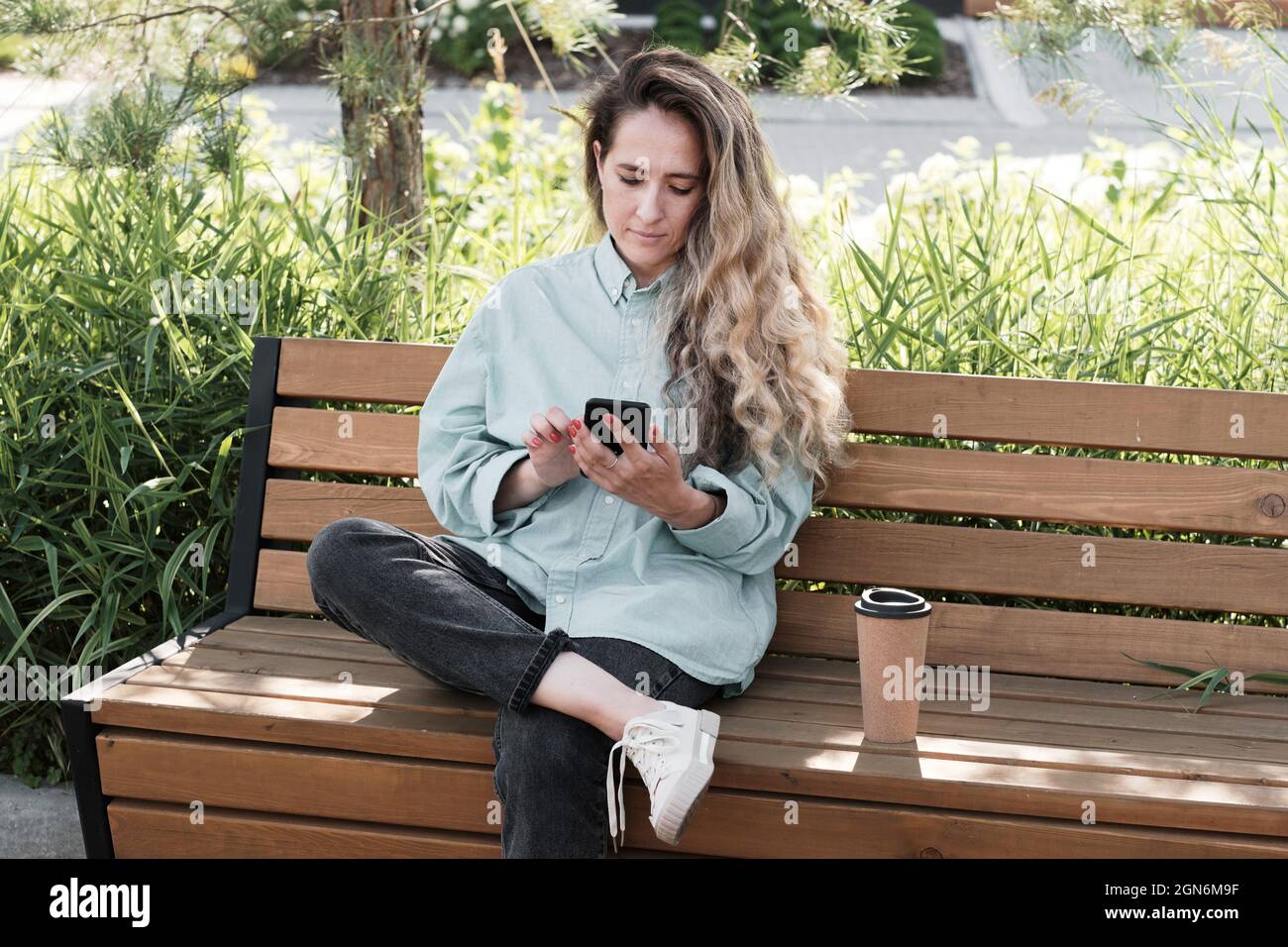 Horizontal mittel lang Porträt von attraktiven kaukasischen Frau mit langen Haaren tragen Casual Outfit sitzen auf Holzbank im Freien mit Smartphone Stockfoto