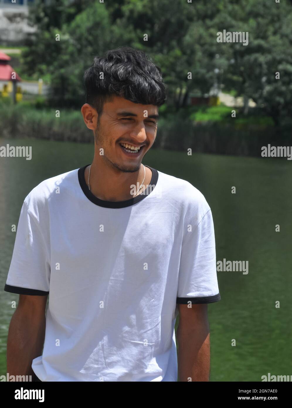 Porträtaufnahme eines jungen indischen Mannes, der neben dem See mit einem weißen T-Shirt steht und lächelt Stockfoto