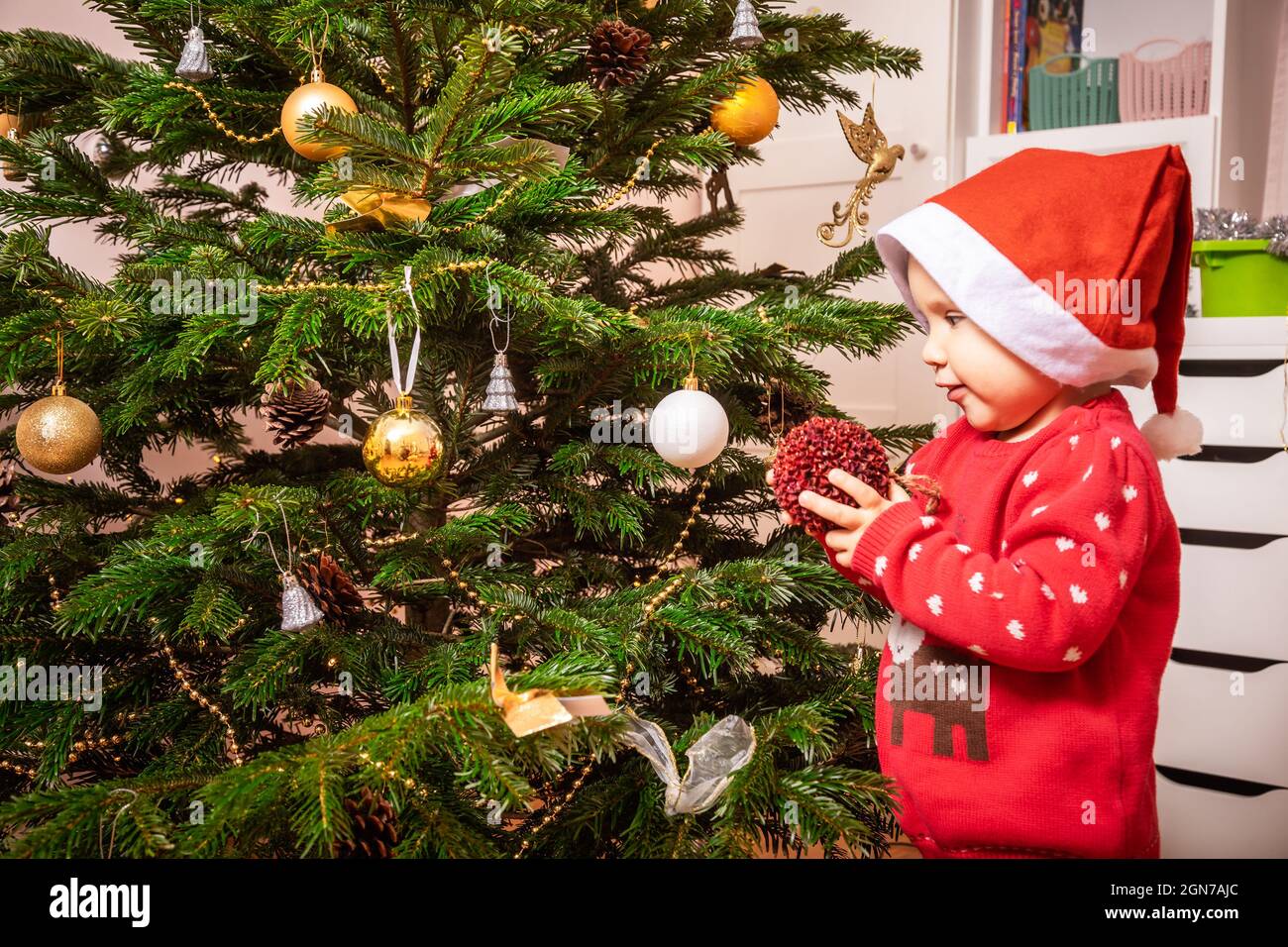 Kleines Mädchen trägt roten weihnachtspullover hilft, Weihnachtsbaum zu Hause zu schmücken Stockfoto
